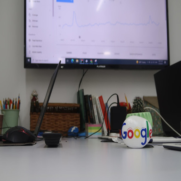 A desk with laptops, a monitor showing analytics, and a Google-branded planter. Ecommerce marketing company workspace.